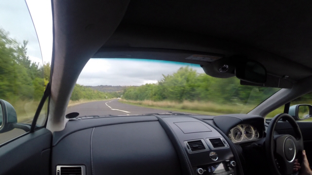 ARE THESE POOR LANE MARKINGS? - Page 1 - Advanced Driving - PistonHeads - The image is a photograph taken from the perspective of a vehicle passenger. The dashboard is visible, showing an integrated navigation system at the center, flanked by air vent controls on either side. The front of the dashboard houses a central console with various buttons and displays, typical of modern automobiles.

The car features a black leather interior, with the vista dominated by a tree-lined road that curves to the right, leading into a landscape that appears to be a mix of open fields and wooded areas, with a hint of blue sky indicating the start of daylight. There is a reflection in the car's side view mirror, giving a glimpse of the interior where fingertips can be seen holding onto something.

The image captures a sense of motion, as suggested by the speedometer display in the car, and the uneven reflection on the side window suggests movement on the road. The photograph appears to be turning right, as indicated by the angle at which the side mirror is positioned, held secure by the hand shown only partially in the reflection.

The scene conveys a sense of journey, with the interior of the vehicle inviting, and the scenery on the road outside indicating travel through a rural or semi-