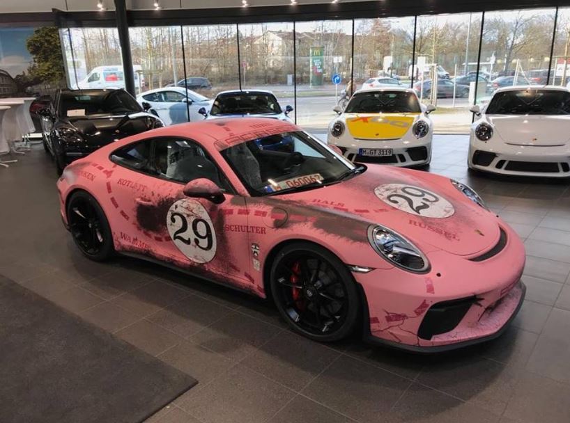 GT3 Touring - Page 42 - 911/Carrera GT - PistonHeads - The image showcases a collection of Porsche cars in a showroom setting. The main focus is a front view of a vibrant pink Porsche car, with the number 29 prominently displayed on the side. This car stands out with its custom spray paint job. The showroom has a sleek, modern design with large windows revealing other cars and cars outside in a landscaped area.