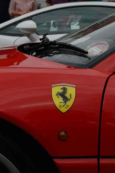 A red car is parked in a parking lot - Pistonheads - The image showcases a red sports car parked outdoors, its nose pointing to the right. A distinctive yellow Ferrari badge is prominently displayed on the front of the car, indicating its brand. A small section of a white car can be seen in the background, suggesting that the red car is parked in front of it. Furthermore, a person's hand is visible in the frame, possibly a photographer or a passerby.