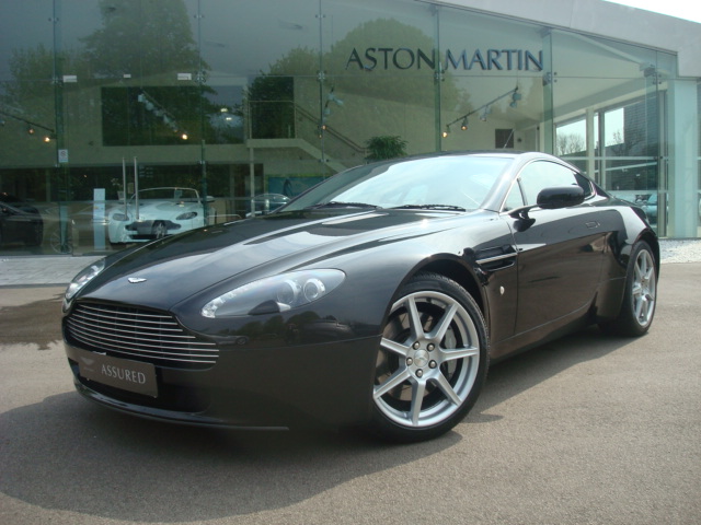 Pistonheads Gang - The image shows a sleek and shiny sports car parked in front of a showroom with large windows. The car is predominantly black with a shiny finish, reflecting some of the showroom's lights. The vehicle features a low profile, prominent angular lines, and distinctive round wheels that are likely to be at least 20 inch rims, suggesting it's a high-end luxury or sports car. The type of the car isn't definitively clear from the image, but it resembles a DBS or Greenfield styled Aston Martin, both of which are known for their sporty and aerodynamic design. The showroom has a modern and clean architectural style, with a large reflective window to the left and a glass door with metal accents to the right. Another car is visible in the reflection of the shop window. The sky is blue and clear, and the ground is a light stone color.