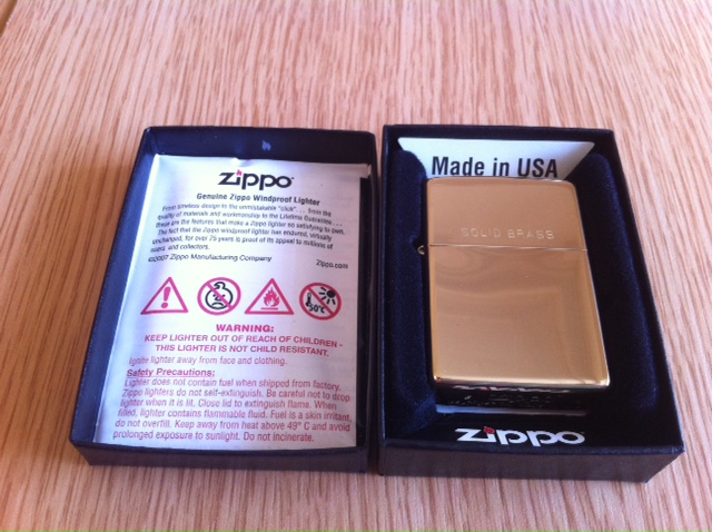 Show us your Zippo... - Page 7 - The Lounge - PistonHeads - The image shows a shiny gold lighter in a black box on a wooden surface. The lighter and its packaging are positioned closely together, creating an interesting contrast between the luminous gold of the lighter and the black of the packaging and the surface underneath. Adjacent to the closed box is an open one, displaying a warning sticker for safety precautions like keeping away from children and not smoking near children. The text on the sticker is clear and legible, suggesting it is part of the product's official or national safety protocols. The overall scene suggests a product that comes with care instructions for consumer safety.