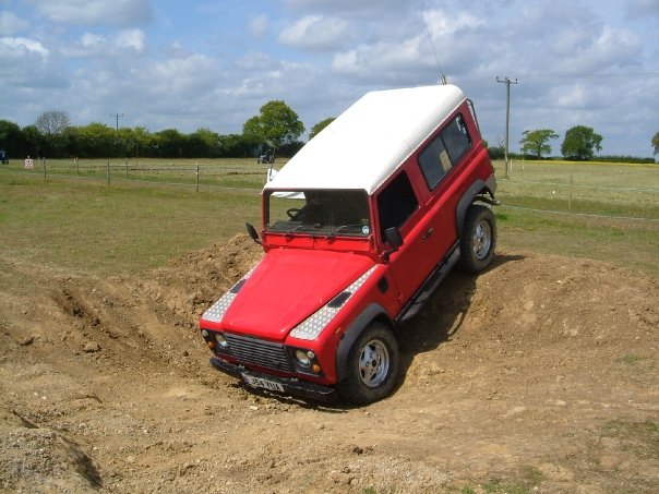 Pistonheads - The image depicts an adventurous scene in an off-road setting. A **white and red** **sports utility vehicle (SUV)** is tilted at an extreme angle on its side in a muddy area, adding a sense of action and excitement to the image. It appears to be a dirt track, suggesting that the SUV might be involved in an off-road driving activity. The sky is filled with **clouds**, suggesting that the weather might be overcast on that day. Given the unusual position of the SUV, there may be a level of skill or skillfulness involved. The background is relatively sparse, with no significant landmarks or features visible, keeping the focus on the adventurous vehicle and the track it's navigating.