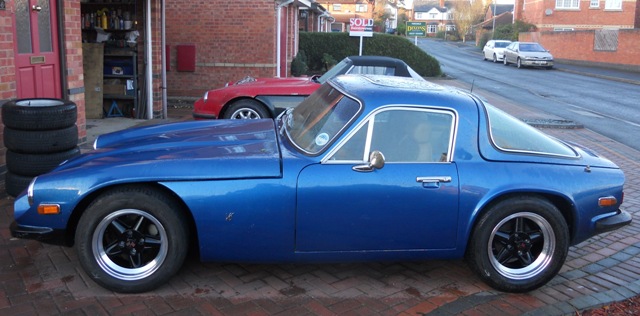 7/16" wheel studs - Page 1 - Classics - PistonHeads - The image shows two classic cars parked on a brick driveway. The foreground features a vibrant blue convertible with its roof down, parked adjacent to a garage. In the background, a red sports car is parked on the grass behind a bush, facing the brickground direction. The setting suggests a residential area, with houses visible in the background. The perspective of the photo is slightly from the side, providing a view of both cars from a slight angle, highlighting the classic design elements of both vehicles. The bricks beneath the convertible are arranged in a diagonal pattern.