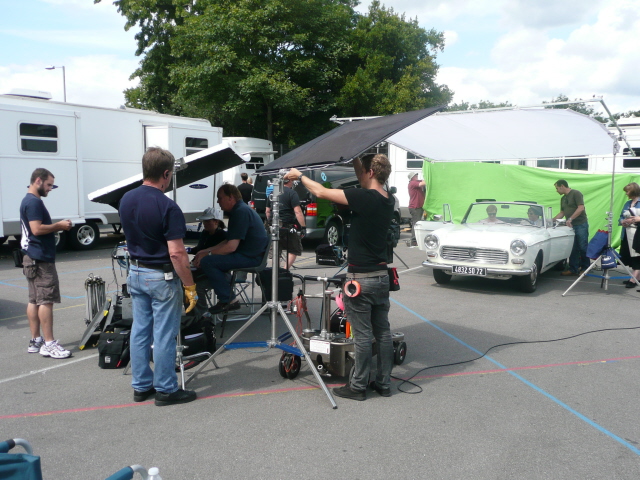 Anyone own a well known car from a tv series or film? - Page 4 - Classic Cars and Yesterday's Heroes - PistonHeads - In this outdoor setting, there's a sense of organized chaos. A white car is the center of attention, as a group of people perform various activities around it. One individual is adjusting what appears to be a large tent or umbrella, ensuring that it provides adequate shade for the adjacent chairs. Another person stands nearby, perhaps overseeing the proceedings. The environment is dotted with multiple vehicles and stone buildings painted in shades of white and blue, adding to the pleasant ambiance. The scene is filled with life, with individuals scattered around, each focused on their own tasks.