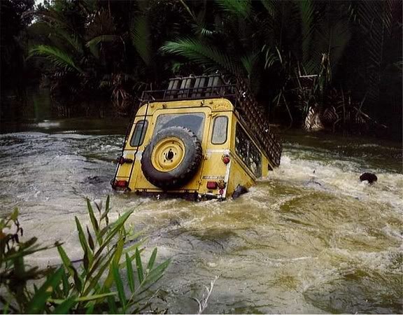 Defender making an entry. - Page 1 - Off Road - PistonHeads - In the image, a large, yellow camping truck is suspended within a river. The truck is partially submerged, with the water level reaching up to its windows. Various equipment, including bicycles, is secured on the top of the truck, suggesting that it might be on an adventure or expedition. The surroundings are lush green, indicating that the truck is in a natural, possibly jungle-like, environment. The scene appears to be choppy and turbulent, indicating that the water level is rising rapidly.