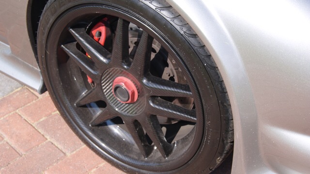 New gto3 owner - Page 1 - Noble - PistonHeads - The image shows a close-up view of a car wheel. The wheel appears to be made of a black material with a red center. It features a five-spoke design with a large bolt extending from the center, which is likely the hub for fastening to the hub nuts on the wheel studs. The wheel is mounted to the inside of a vehicle with part of the car fender or door panel visible in the right side of the image. The background displays a sidewalk made of brick pavers. There is no visible text or branding on the wheel or in the immediate vicinity.