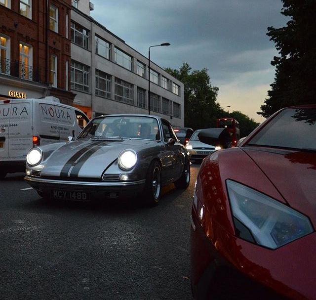 The Beautiful Porsche Picture - Page 5 - Porsche General - PistonHeads - The image shows a scene at dusk with vehicles on a road. There's a red sports car in the foreground and a white delivery van in the background, both appearing to be in motion. In between these two, there's a silver sports car that seems to be in motion as well; it has a distinctive stripe down its middle. The street is illuminated by the headlights of the vehicles and some residual light in the sky, suggesting that it's the evening or early nighttime. The architecture of the buildings on either side of the road suggests a denser urban setting, possibly a city or town center.