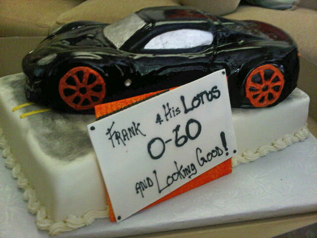 My 40th Birthday Cake - Page 1 - Elise/Exige/Europa/340R - PistonHeads - This image features a round, coated cake decorated to resemble a black sports car with orange flames. The cake is placed on a table, and a white sign reading "Frank 4 His Lonos" with a message of encouragement is placed beside it. Behind the cake, there are more objects on the table, including a couch and additional cake decorations.