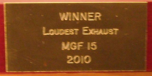 Pistonheads - The image features a plaque with embossed text on a wooden background. The plaque is predominantly gold in color with the text displaying a 'WINNER' title on top followed by the words 'LOUDEST EXHAUST'. Below that, there is a meaningful notation that indicates 'MGF 15 2010'. The text is clear and prominently displayed on the plaque. The background provides a nice contrast to the gold text, making the text easily readable for viewers.