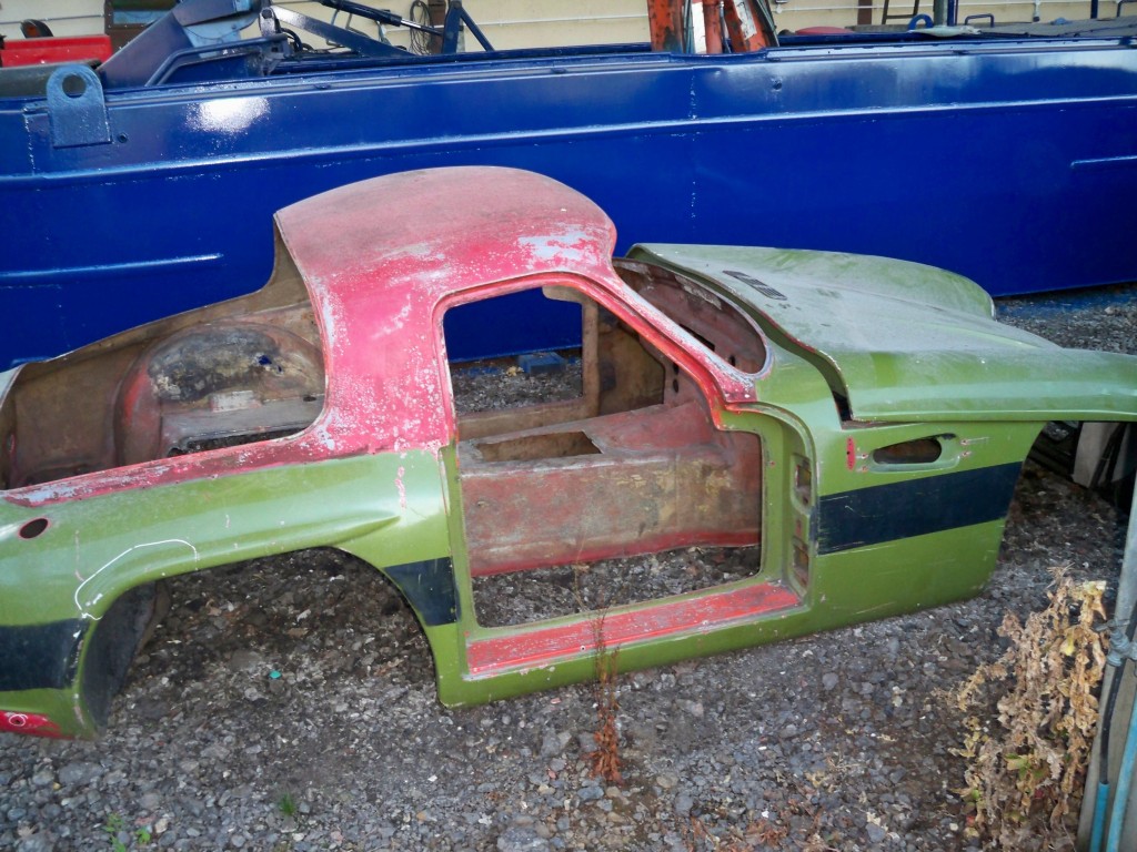 Early TVR Pictures - Page 104 - Classics - PistonHeads - This image showcases the front view of a vintage car that appears to have some wear and tear. The car's front hood is open, revealing its engine area, which is covered in pink paint. The front car fender is also red and has a green stripe adding to the colorful appearance. The vehicle is situated on broken glass and a gravel-like surface, suggesting it might be discarded or in the process of restoration. The car itself is positioned next to a blue vehicle, possibly indicating a workshop or storage area.