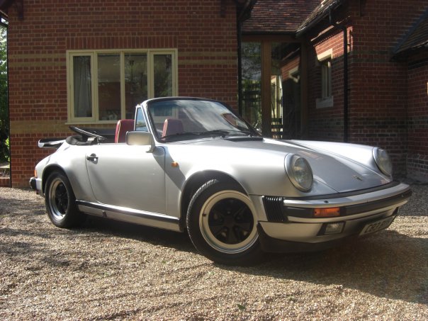 Porsche 911 3.2 - Page 1 - Readers' Cars - PistonHeads - The image features a silver Porsche 911 parked on a gravel driveway in front of a house. The car is positioned at an angle to the camera, allowing for a clear view of its front end. The Porsche's prominent, round headlights and the sunlit reflection of the building on its surface are striking features. The scene conveys a calm and serene atmosphere typical of a residential setting.