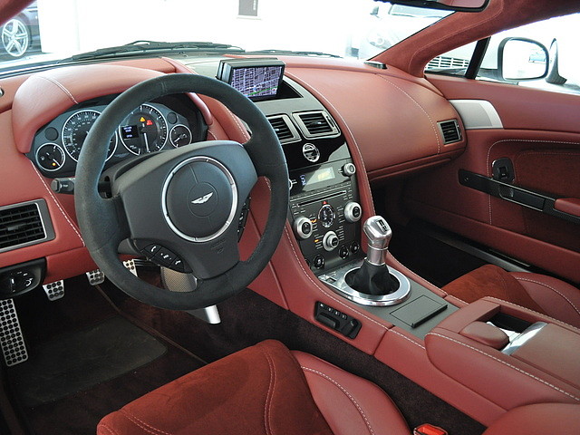 window tints - Page 1 - Aston Martin - PistonHeads - The image presents the interior of a luxurious car. The car features a dark red interior with a large infotainment screen centered on the dashboard. Behind the steering wheel, there is a rectangular navigation display. The steering wheel itself is adorned with a logo, suggesting the car brand. The dashboard has a high-gloss finish and the instrument cluster is clearly visible.