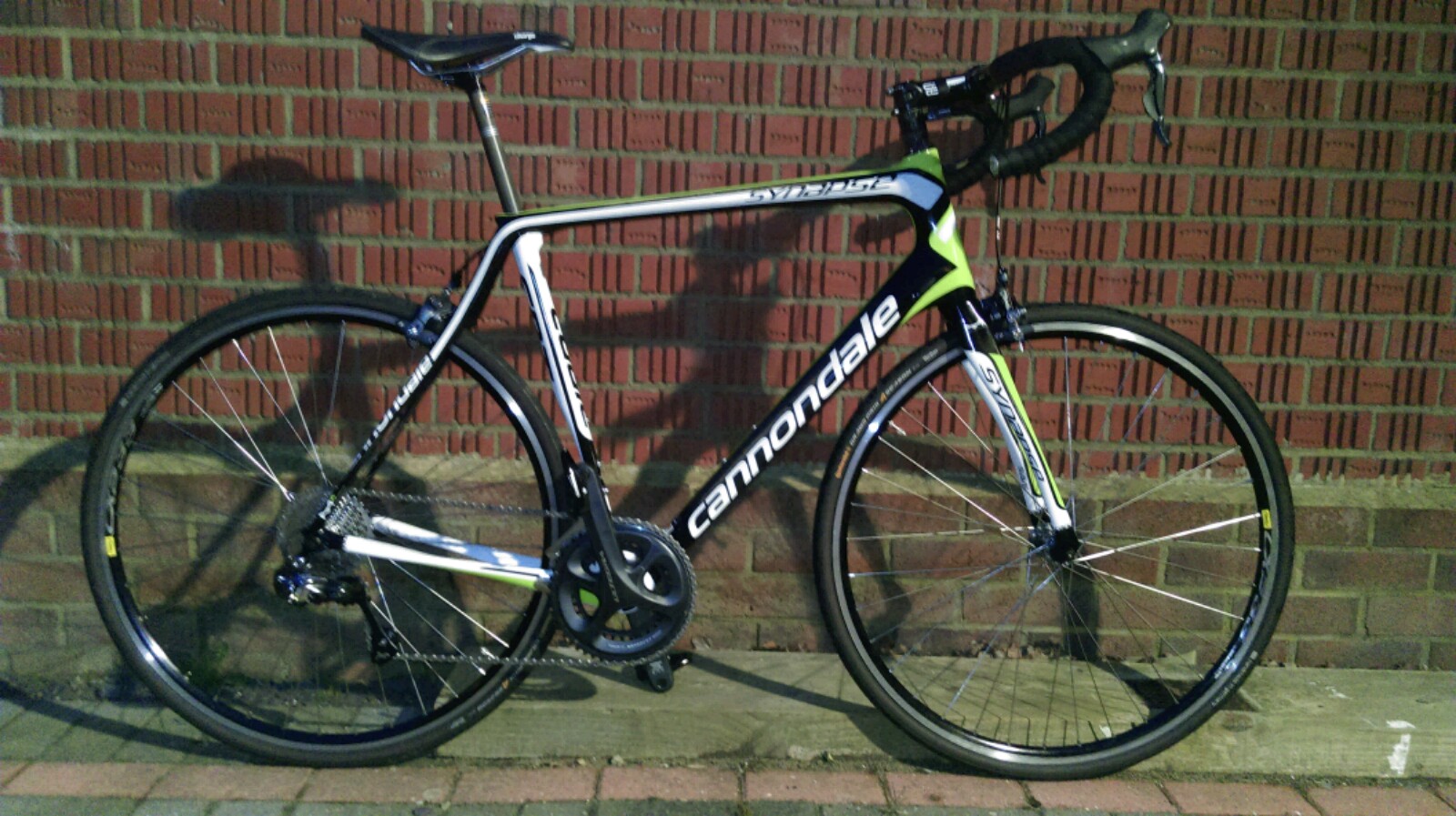 A bike parked on the side of a road - Pistonheads - This image features a modern road bike parked on a paved sidewalk. The bike has a sleek frame and is equipped with multiple gears, signaling it is designed for a range of terrains. The bike's color scheme is primarily black and white, with green accents and the word "CAMPAGNER" visible on the chainset. The bike is leaning slightly, and there is a red brick wall visible behind it, creating a contrast between the urban environment and the bike itself.