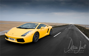 Pistonheads Lambo Locations Photoshoot - The image depicts a yellow sports car driving on an open desert road. The road is bordered by a minor expanse of greenish vegetation on the right side of the vehicle. The vehicle appears to be moving at a fast pace, evidenced by the blurred backdrop, which also includes a subtle road marker and a hazy sky with clouds. The image conveys a sense of speed and isolation, emphasizing the car's solitary journey through the vast desert landscape.