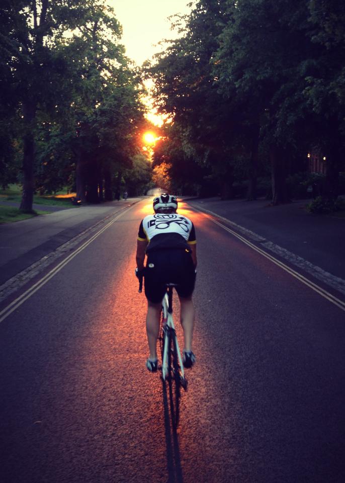 The "Photos From Today's Ride" thread... - Page 133 - Pedal Powered - PistonHeads - The image depicts a daring sunrise bicycle ride down a country road. A cyclist, clad in a black and white uniform with a large pattern that resembles a sheep herd, is crossing a highway with a backdrop of the sunrise, casting a radiant glow over the scene. The road is lined with lush green trees on both sides, reflecting the cyclist's path in their side mirror. The cyclist's reflection on a glistening wet road ahead of him adds a sense of motion to the serene setting.