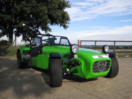 Looking for a Superlight R300 - Page 1 - Caterham - PistonHeads - The image displays a vibrant green vintage racing car parked on a gravel surface. It has distinctive grilles on the sides and prominent headlamps casting a light blue hue on the dusty ground. The vehicle features a convertible top and wide, black, oversized wheels that add to its retro charm. It appears to be a two-door car with the door on the left, and there's a white number 7 on the front grille. The sky is slightly overcast with some clouds, suggesting an outdoor setting during either sunrise or sunset, as the light is soft and diffused.