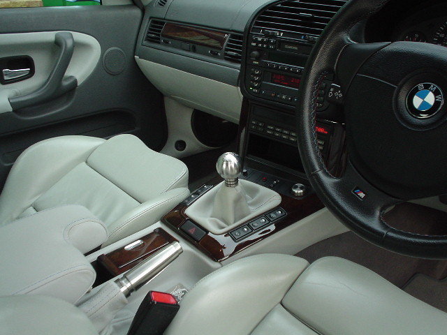 My 1997 E36 328i - Page 1 - Readers' Cars - PistonHeads - The image depicts the interior of a car, featuring the driver's side perspective. A luxurious, black leather steering wheel is positioned on the right. In the center console, there's a large ashtray with a cigarette lighter on top. The car's dashboard and the gear shift lever are also visible, with various buttons and indicators. The upholstery of the car seats is white in color.