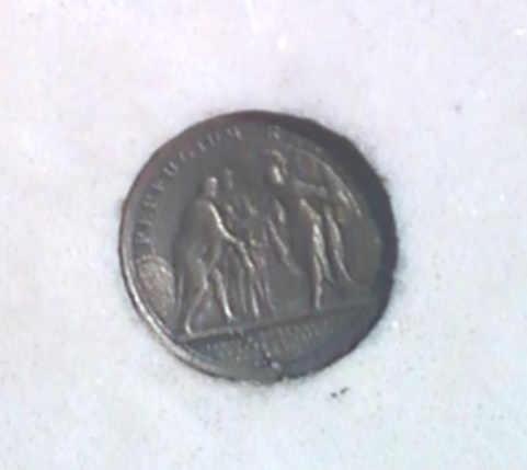 The image shows a metal coin with embossed details resting on a snowy surface. The coin features a relief illustration of a seated figure, likely a classical or historical figure, holding a torch with both hands. There is text on the coin, including what appears to be the script of a coin inscription, which is commonly Latin. The coin's metals and the texture of the surface indicate it's well-used or possibly it is a counterfeit. The overall scene suggests a snowy, outdoor setting.