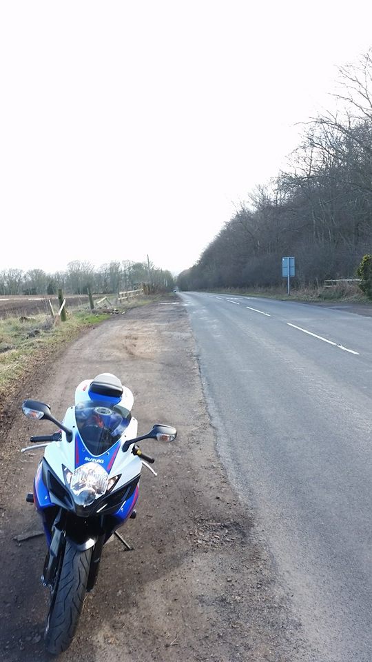 A motorcycle parked on the side of a road - Pistonheads - The image captures a tranquil rural road in daylight. A single motorcycle, with its blue, red, and white colors, stands out against the quiet and empty road. The motorcycle is parked on the right side of the blacktop, with its visible rear angle. The road stretches out into the distance, lined on both sides with trees, under a grayish sky that resembles a cloudy day. Beside the road, closer to the camera, there is a small, uncultivated patch of earth, hinting at solitude or the off-season.