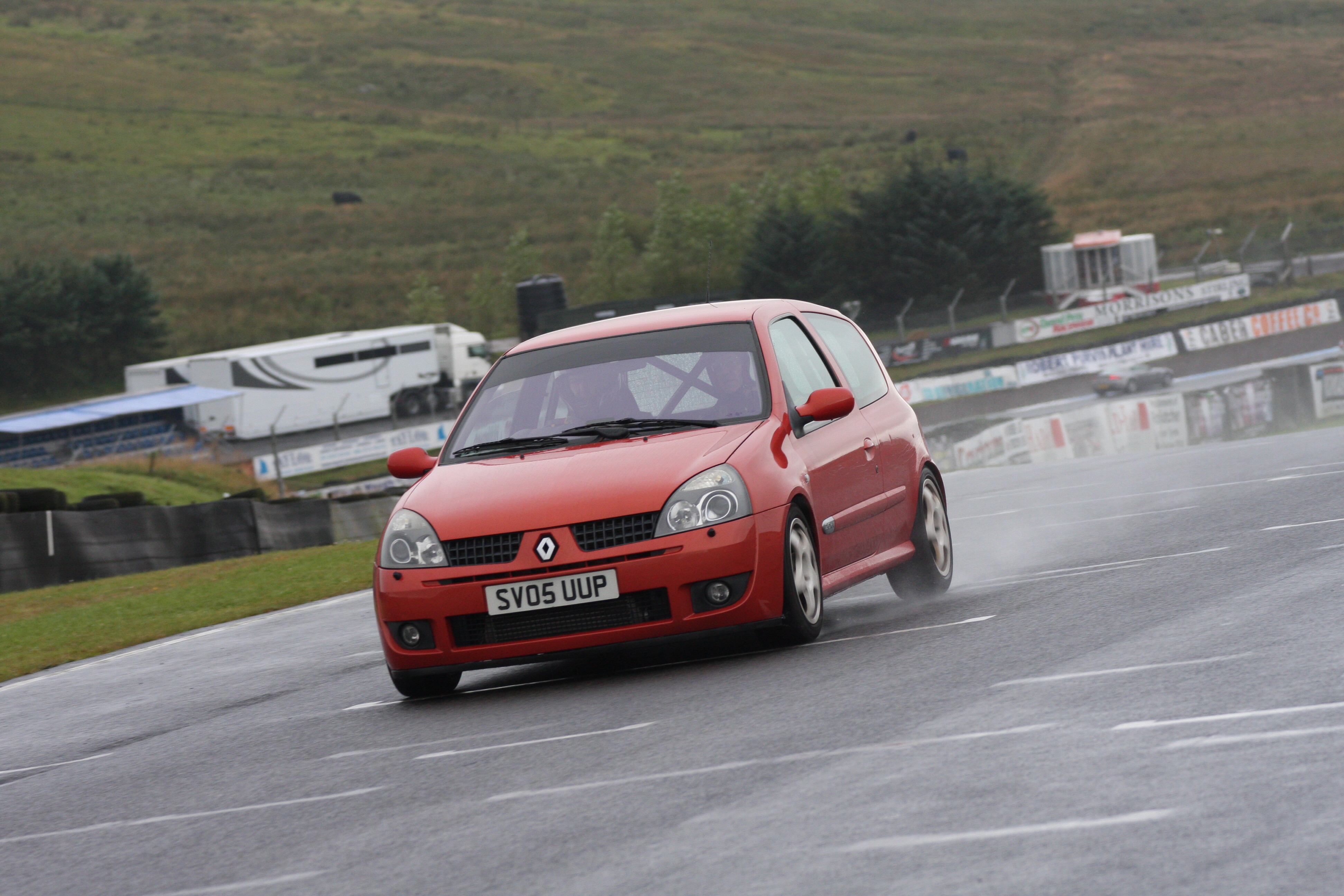 What is your track day car, and why?  - Page 10 - Track Days - PistonHeads - The image depicts a red Renault Clio hatchback car in motion on a race track. The car appears to be a racer, given its aerodynamic design and the dark-tinted windows. The car has black wheels and is leaning to one side, suggesting high speed or a sharp turn on the track. In the background, there are other vehicles, possibly racing or parked, and a series of winding roads or tracks. The setting appears to be a rural outdoor area, as suggested by the grass and the varying shades of green that can be seen in the environment.