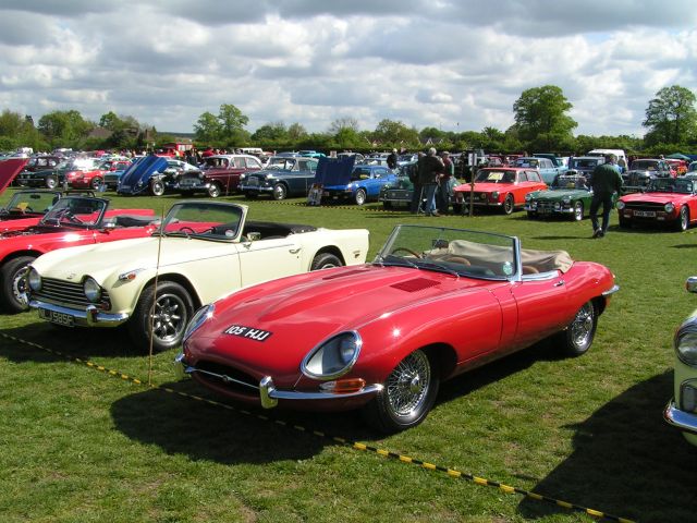 Pistonheads - In the image, a vibrant outdoor car show is taking place on a grassy field under a partly cloudy sky. The central focus is a striking red convertible parked in the foreground, with other classic cars artfully arranged around it. The vehicles exhibit a spectrum of colors ranging from pristine white to deep red and earthy greens. People can be seen mingling among the cars, adding a lively atmosphere to the event. The scene radiates a sense of nostalgia and automotive appreciation, capturing the essence of a classic car rally.