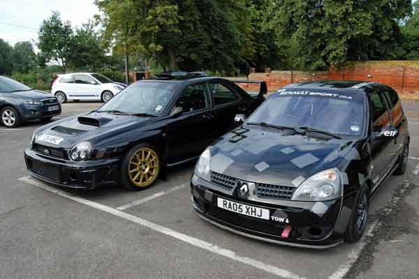 A car that is parked next to a parking meter - Pistonheads - The image shows a meticulously maintained black Renault Sport hatch parked next to a black sports car in what appears to be a parking lot. Both vehicles are customized with various decals and finishes, and the Renault has gold-colored rims and a black body kit that enhances its sporty look. On the back of the Renault, there is visible branding for Renault and the Renault Sport series. The sports car has a unique grill design and is also labeled with "Renault Sport" and other logos. The setting includes other cars parked in the background, indicating a common area for vehicle parking.