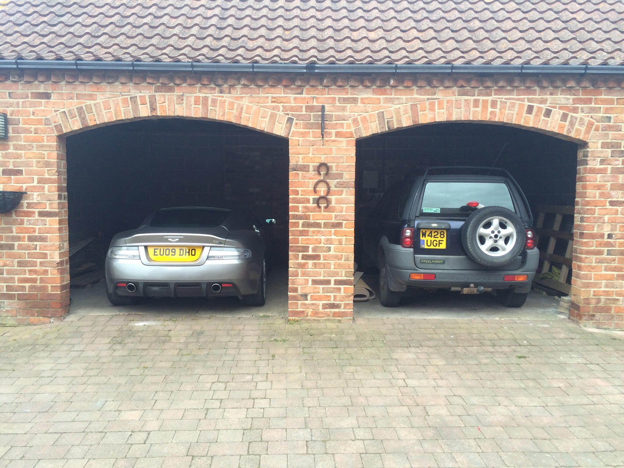 A car parked in front of a building - Pistonheads - The image captures a scene of two cars parked under a brick archway of a driveway. The car on the left is a sleek, silver sports car with a license plate reading "S00 ALU". The car on the right is a robust black SUV with a license plate featuring "UYG". Both cars are parked neatly side by side. The archway has a door on each side and is made of red bricks, adding a rustic touch to the scene. The driveway appears to be constructed of gray stone, extending into the distance where it meets the rest of the street.