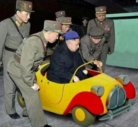 North Korea photoshop contest - Page 10 - The Lounge - PistonHeads - The image shows a group of men in military uniforms, featuring hat-like headgear and one with a striped tie. They are standing around a small car that appears to be a part of a road parade or military exercise. The car is a compact design with solid coloring in yellow and red, featuring chrome accents and shuttered wheels. There is a champagne bottle resting in the tiny rear space. The men are engaged in a discussion or observation, possibly strategizing or monitoring the situation.