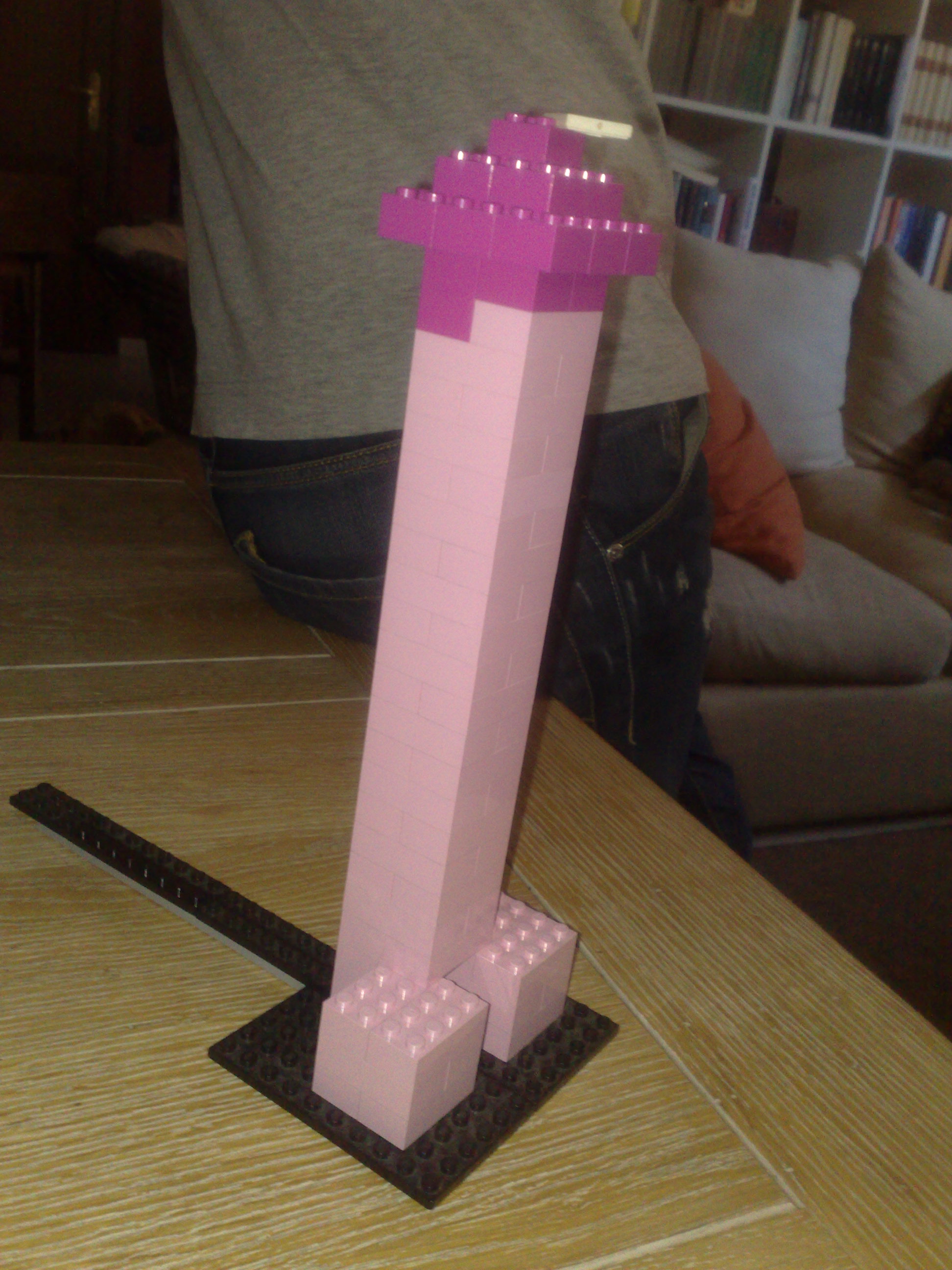 The image captures a creative and artistic display on a table. The centerpiece is a Lego structure that takes the form of a two-legged tower. The color scheme is striking, contrasting a pink base with a tall purple core.

The Lego structure's upward and outward expansion gives it a modern and abstract look. It stands out against the more traditional background.

In the blurred background, the contrast is further emphasized with the figures of people, who appear to be adults. Their figures provide a sense of scale to the image.

The setting seems to be a living room or a similar domestic space, suggested by the presence of a couch and a bookcase. The room feels warmly lit, possibly by natural light from outside the image.