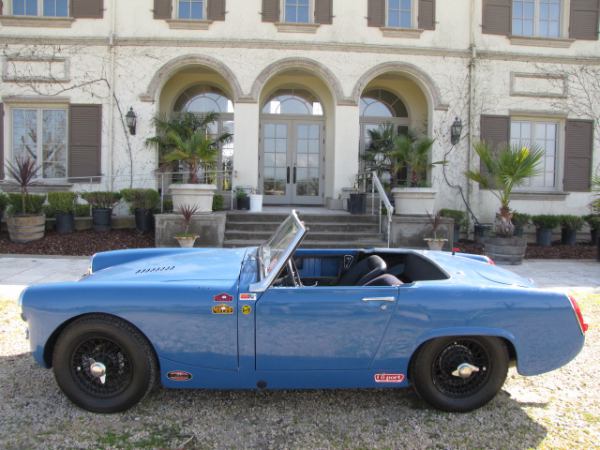 Modified Midgets - Page 1 - MG - PistonHeads - The image showcases a striking blue convertible parked on a gravel lot in front of a two-story house. The car features a distinctive grill and white-walled rims, giving it a vintage flair. The house itself is large with a stately entrance, decorated with black gates and palm trees lining the driveway. The scene suggests a warm, sunny day, ideal for a leisurely drive in the convertible. The overall composition conveys a sense of comfort and luxury.