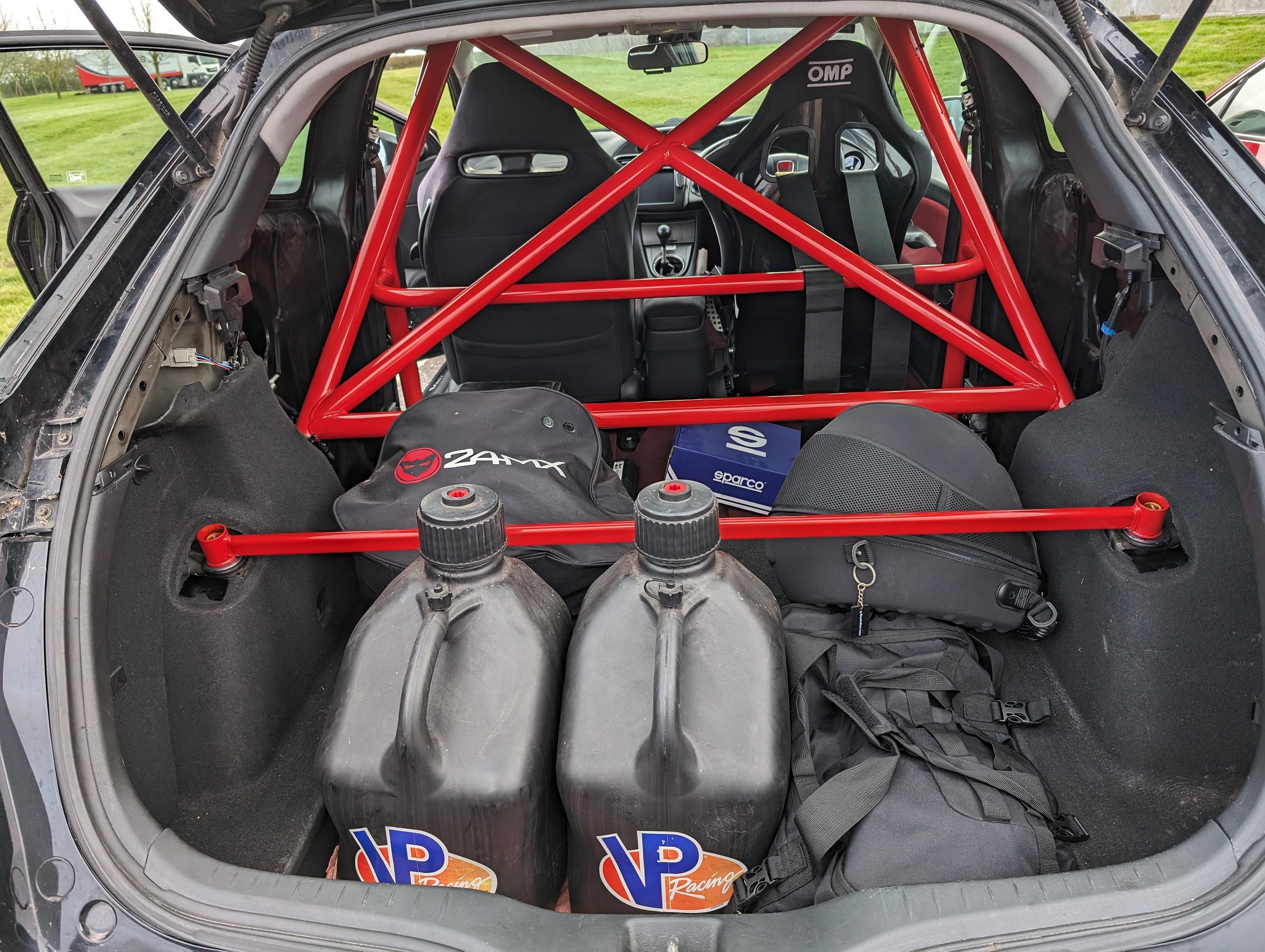 Pistonheads - The image depicts the interior of a car, viewed from the perspective of someone standing outside. It appears to be a rally car or race car, as evidenced by the roll cage and harnesses in the front seat. There is a red and white car chassis visible, with various equipment parts such as a jack and tools scattered around. On the backseat, there's a set of suitcases and other travel essentials, suggesting that the car might be used for competition or traveling purposes. The image has a watermark in the bottom right corner, indicating it may have been edited or manipulated.