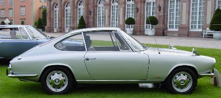 Identify Pistonheads - This image depicts two classic cars parked on a lawn in front of a large, red brick building with many arched windows. The car in the foreground is a light-colored convertible with the top down, showcasing its interior. The second car, located behind the first, is a darker-colored sedan with similar vintage styling. There are well-manicured hedges in the background, adding to the old-world charm of the setting. The presence of the potted plants and the orderly arrangement of the cars suggest this might be a special event or a display of vintage cars.