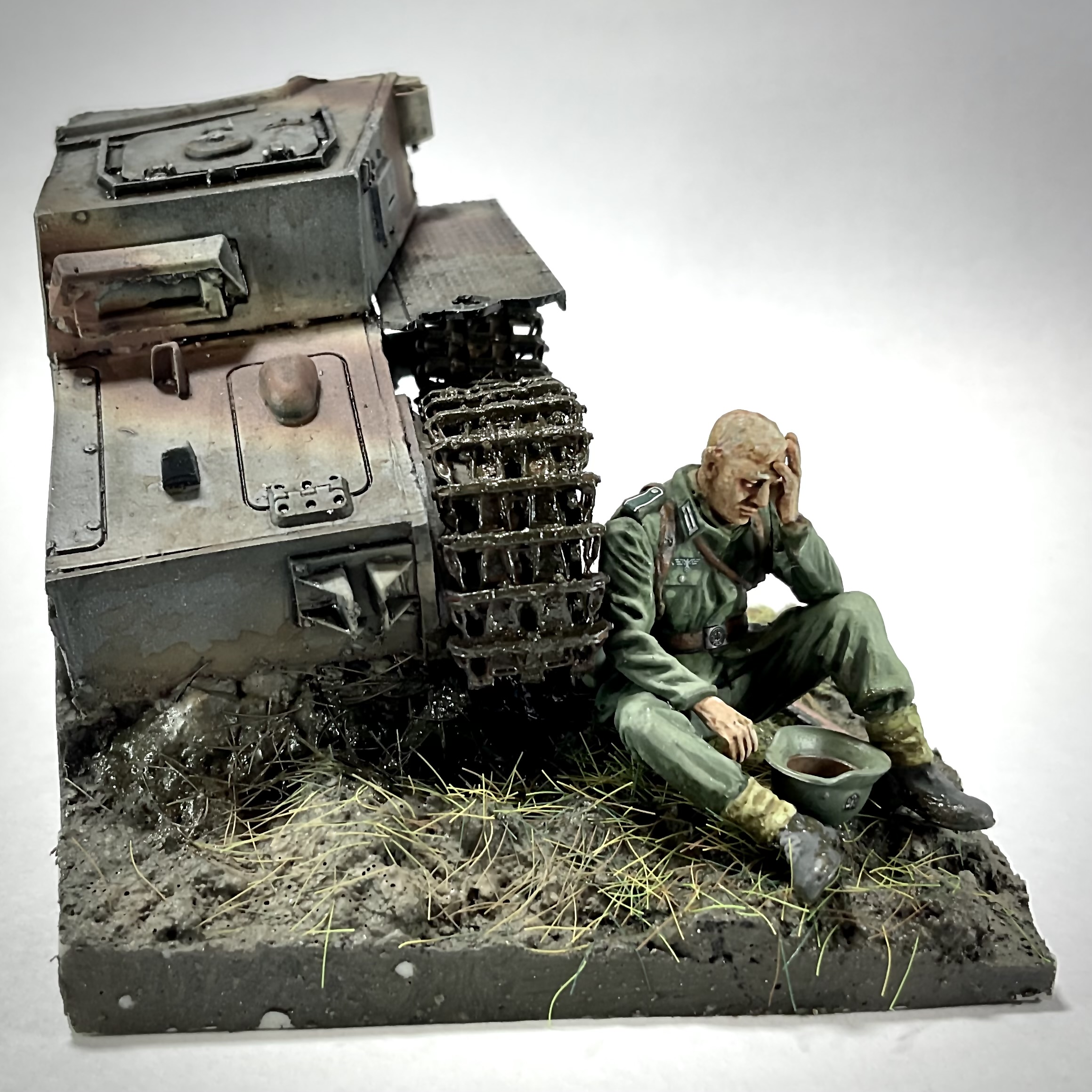 Pistonheads - The image displays a miniature battle scene with a tank and an infantryman. A soldier in military fatigues, complete with boots and a helmet, is kneeling on the ground next to a tank that appears to be damaged or abandoned. He is holding his head, suggesting distress or defeat. Both figures are positioned under a canopy of what might be a tarp or a piece of cloth, providing shelter. The background is not detailed, but it gives an impression of a battlefield environment. The entire scene is presented on a small table or shelf. The image has a realistic and detailed aesthetic, typical of a model diorama, and captures the poignant moment in a way that suggests narrative and storytelling.