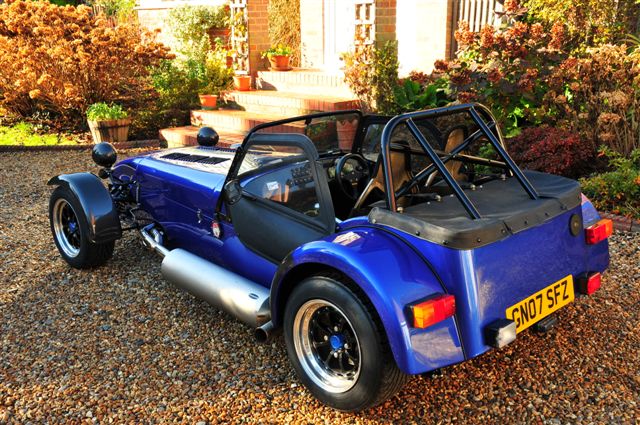 Xmas Pistonheads Merry - The image showcases a blue three-wheeled car, predominantly occupying the frame and set against a backdrop of a gravel driveway and flowering trees. The car has a distinct vintage look with its small size and black wheel rims. It also has a large windshield and a set of two wheels at the front and the other at the rear. A small antenna is visible on top of the car.