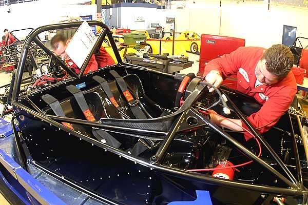 A man standing in front of an open suitcase - Pistonheads - The image depicts an indoor setting where a person is working on a silver racecar. The individual is dressed in a red top, giving a sense of team coloration, and appears to be focused on the vehicle. The racecar is open with its roof off, revealing the driver's area and chassis inside. The surrounding environment seems to be a workshop or garage, with tools and equipment visible in the background.