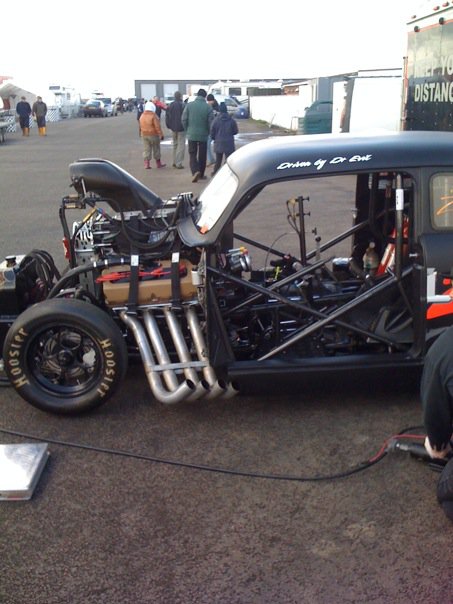 What did you do in the garage yesterday? - Page 5 - Chimaera - PistonHeads - In this image, a black drag racing car is parked on a concrete surface. The car is equipped with several performance-enhancing features including two large rear spoilers, roll cages, and a hood that has been cut out for showcasing the engine. Various wires and hoses are visible, hinting at an intense and well-equipped engineering setup beneath the hood. The drag racing car is surrounded by people, some walking by, others seemingly more engaged, suggesting an event or gathering related to drag racing or vehicle customization.