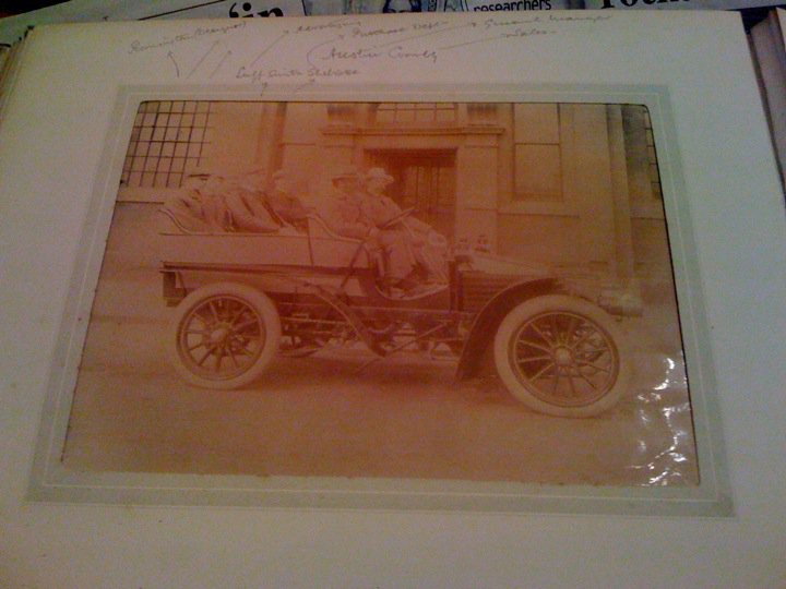 Century Pistonheads Wolesley Turn - The image depicts a vintage black-and-white photograph featuring an early 20th-century car with two passengers. The vehicle is old-fashioned with its design indicating a bygone era. The car is parked on a street with a building in the background, suggesting it's an urban setting. There's a stop sign to the right, which also hints at the context of a public road. The image is slightly overexposed, with some areas brighter than others, and includes handwritten text written on or across the image, which may include names, dates, or descriptions of the photo.