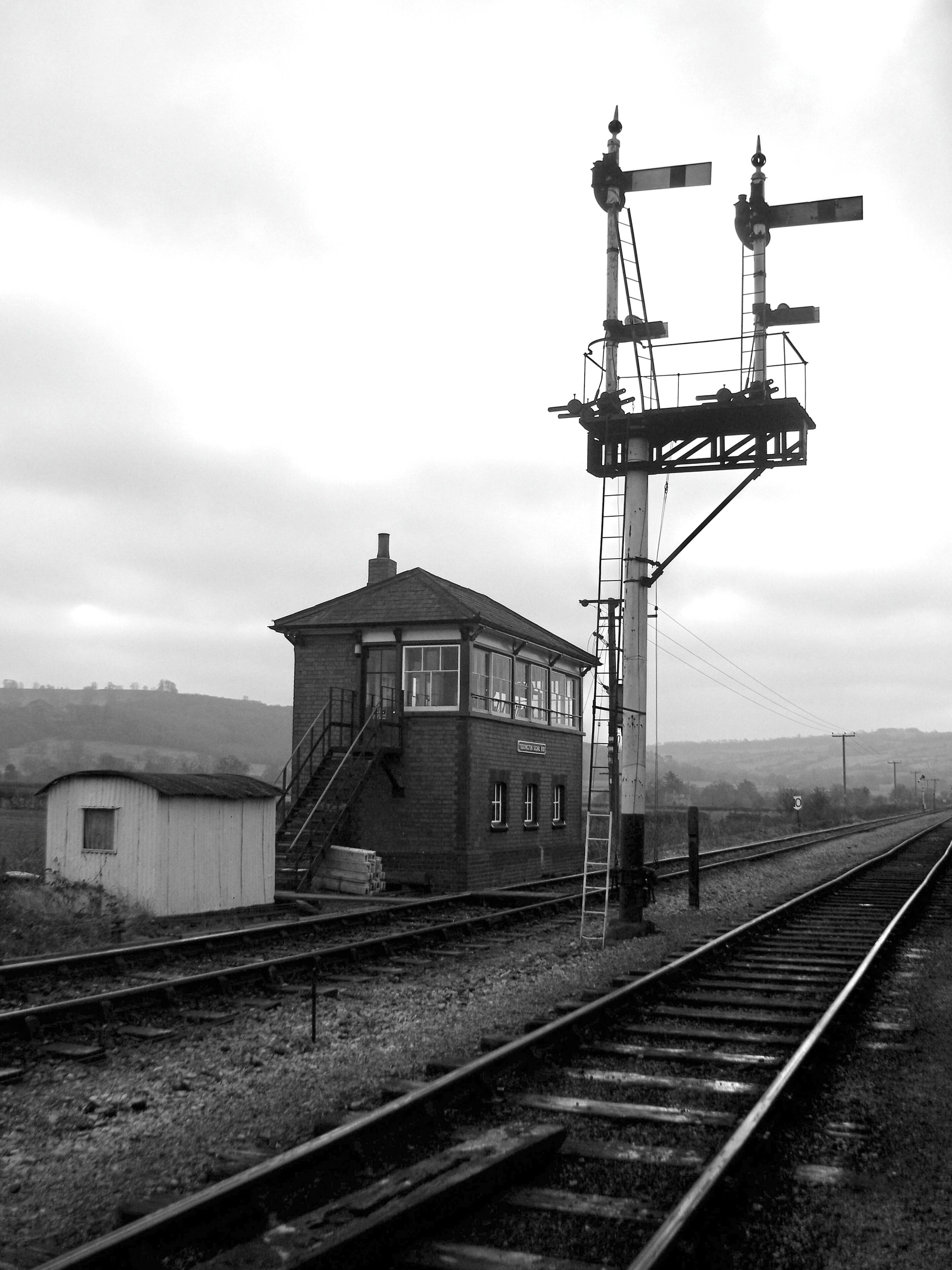 Pistonheads - The image is a black and white photograph depicting an old-fashioned railway scene. A train track with railroad ties stretches across the frame, leading towards a distant train station. On this track, a small signal box with semaphore arms stands at attention. Two railroad workers are positioned inside, attentively observing the tracks and the station ahead. The station itself is simple in design with a single platform visible. The sky overhead is overcast, suggesting an early morning or late afternoon timeframe. A fence encloses the immediate area around the signal box, adding to the vintage feel of the scene.