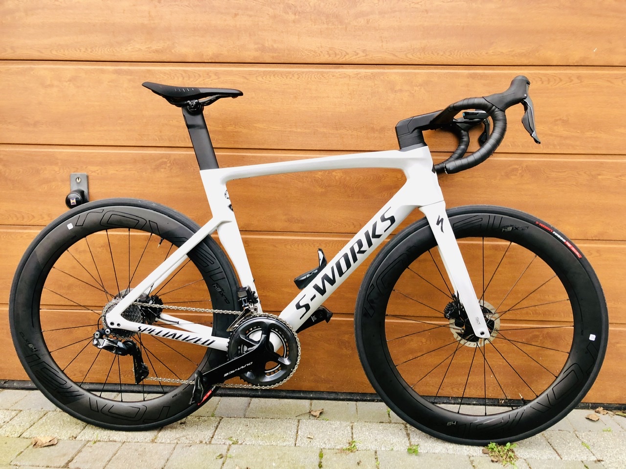 The "what bike bits have you just bought" thread Vol 2 - Page 60 - Pedal Powered - PistonHeads - The image shows a modern road bicycle parked next to a garage door. The bike is white and black, featuring sleek lines and contemporary design elements such as aero seatpost, disc brakes, and wide tires suitable for gravel or long rides. It has several integrated components including a bar-mounted electronic shifting system and hydraulic disc brakes. There are visible brand logos on the bike and the wheels. The background is minimalistic with a wooden garage door and a portion of a house wall.