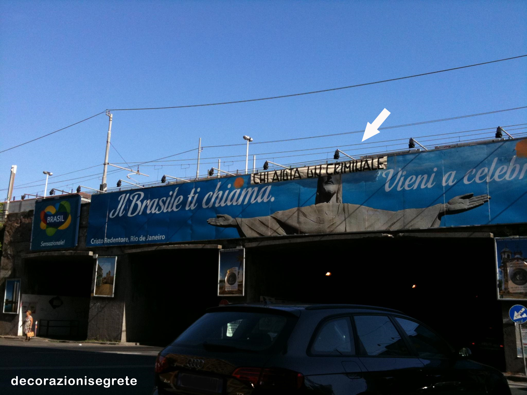 In this image, we see a blue billboard with an illustration of a child hanging from his knees above a Brazilian flag. There is also a sign indicating that the structure is a train station. A black car is parked on the side of the road below the billboard, and a person can be seen walking on the sidewalk. The billboard includes text in Portuguese and Arabic.