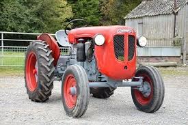 Vixen vs Elan - Page 1 - Classics - PistonHeads - The image shows a single tractor parked on a gravel surface. The tractor is a classic design, with a bright red body and large black wheels. Its cylindrical shape is typical of vintage tractors, and it appears to be in good condition. In the background, there's a dilapidated shed, which gives the scene a rustic and somewhat worn feel. Near the tractor, there's a structure with a metal fence in the foreground, providing context for the setting. The overall composition suggests that this is an old farm or agricultural area.