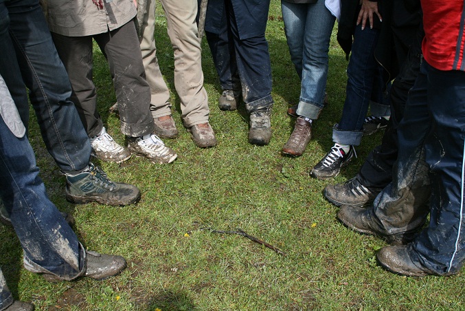 Shoes Dirty Filthy Dirt Mud - The image is a candid outdoor photograph showing a group of people standing closely together, all with muddy shoes and legs indicating they have been in mud or soil. They are arranged in two rows facing each other across a small area of grass. The group appears to be of various genders and ages, suggesting a casual, possibly social gathering that has taken place in outdoor conditions.