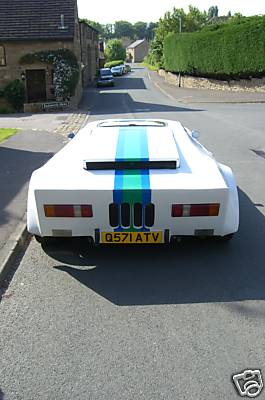 Dutton Pistonheads Proves Universally Hideous - The image shows a **white sports car** parked on a **gray street**. The car features a **green and blue stripe** on the hood, adding a sporty touch to its appearance. The car is positioned in front of a **brown house** with a **white garage door**, suggesting a residential setting. The **sky is clear, indicating a lovely day**. A **posh** upper left-hand corner watermark is present, possibly indicating the source of the image.