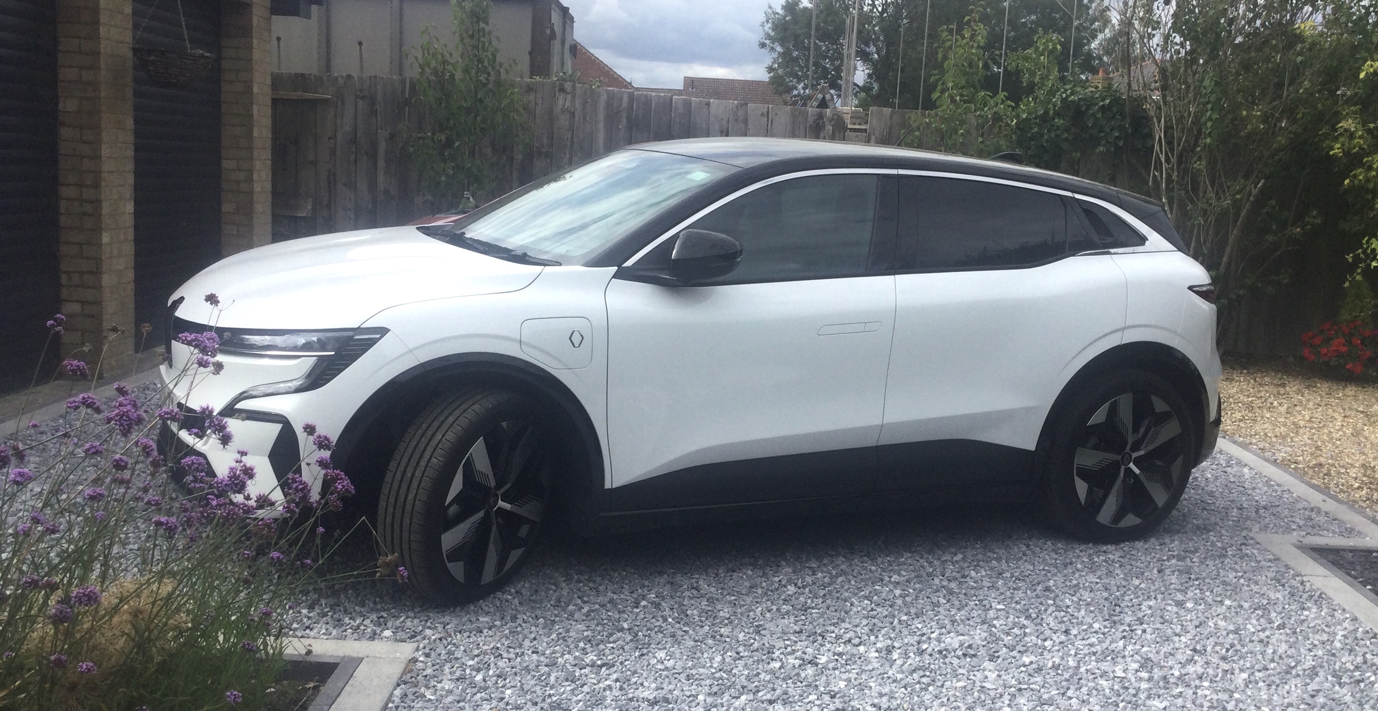 The PH EV photo club - show us your electric cars! - Page 16 - EV and Alternative Fuels - PistonHeads UK - The image features a white SUV parked in front of a house. The car has black accents and is positioned at an angle to the viewer. In the background, there's a well-maintained garden area with plants and a fence, as well as a stone driveway leading to the house. A person can be seen standing near the back of the SUV, partially obscured by the vehicle. The sky is clear, suggesting it could be early morning or late afternoon due to the soft lighting.