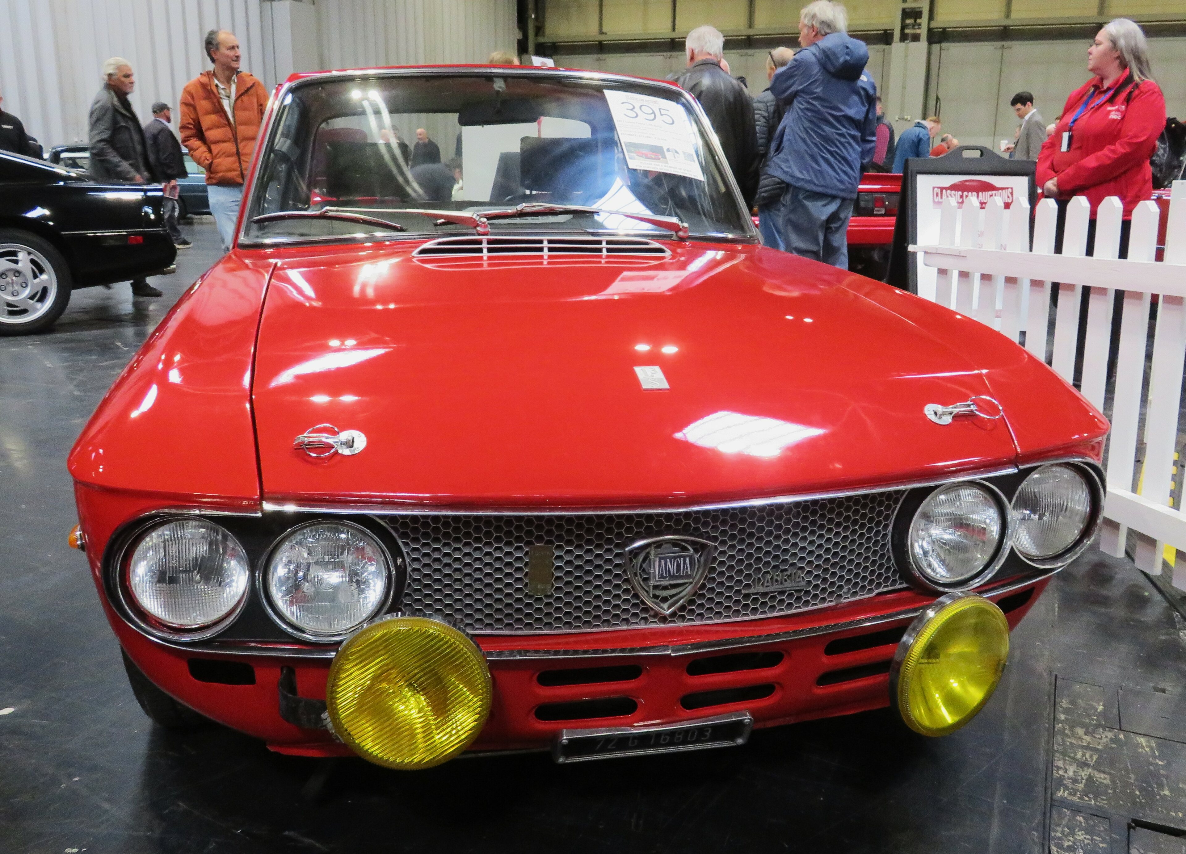 Lancia Fulvia 1.6 HF - Page 5 - Readers' Cars - PistonHeads UK - The image showcases a vibrant red classic car parked in an indoor setting, likely a garage or exhibition area. The car is characterized by its yellow headlights and is adorned with number plates on the front and rear. Its design suggests it might be from the 1970s or a similar era. In the background, there are several other vehicles, including cars and trucks, indicating a collector's or enthusiast's garage. The overall ambiance of the image is nostalgic and filled with automotive charm.