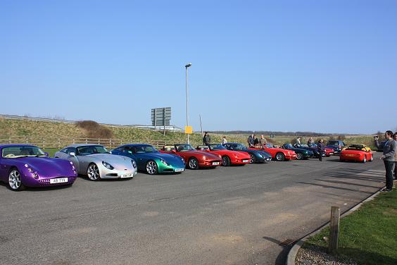 Cobweb Tvrcc Pistonheads Valley Tees - The image showcases a unique gathering on a sunny day. Several vintage racing cars are parked in a row along a dusty road, painted in shades of bright red, cool blue, and poker chips. Each car is distinctively numbered, and the drivers appear to be posing beside their respective vehicles, attracting the attention of a small crowd of onlookers.