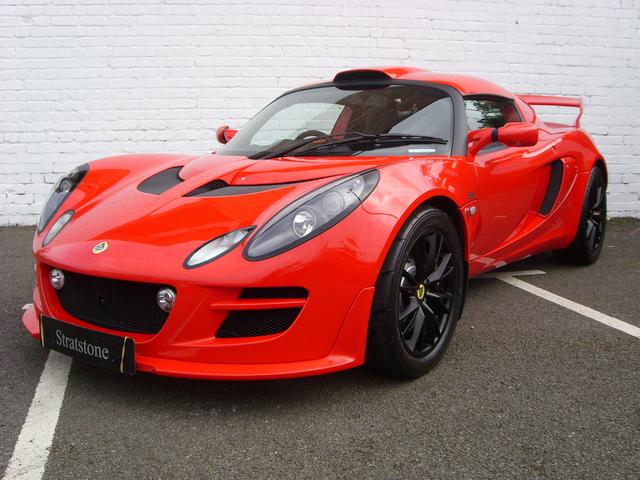 Lotus Exige V6 SC (Exige (S3?) - New Elise SC - Page 7 - Elise/Exige/Europa/340R - PistonHeads - The image shows a sleek, red sports car parked in a parking lot. The car is angled slightly, facing towards the left side of the image, and is positioned perpendicular to a white brick wall. It features distinctive design elements such as large air intakes, aerodynamic bodywork, and three-spoke alloy wheels. The car's vibrant red color stands out against the neutral backdrop of the white brick wall and the road, indicating that it might be a rare or special edition model.