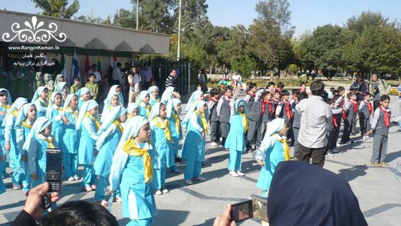 The image depicts a group of children dressed in bright blue traditional clothing, which includes dresses and headscarves, standing in front of a building with trees in the background. Some of the children are seated on the ground, while others are standing, forming a semi-circle. There is a crowd of people to the left of the image, and a flag and benches are visible in the background. The focus of the image appears to be on the children's attire and the arrangement they are in, with the individuals in the crowd and the background elements providing context.