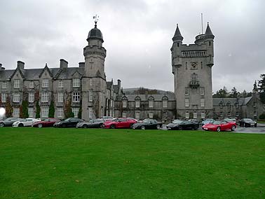 Pistonheads - The image depicts an impressive castle-like structure surrounded by a vast green field. The building stands grandly with three prominent towers and more nuanced turrets. In front of the castle, there's a row of parked cars, painted in a variety of colors, creating a stark contrast against the grass. The sky overhead is cloudy, suggesting an overcast day. Beautifully maintained trees can be seen to the left of the image, adding a touch of nature to the scene. The image seems perfectly framed, capturing the essence of the grand structure and its green expanse.