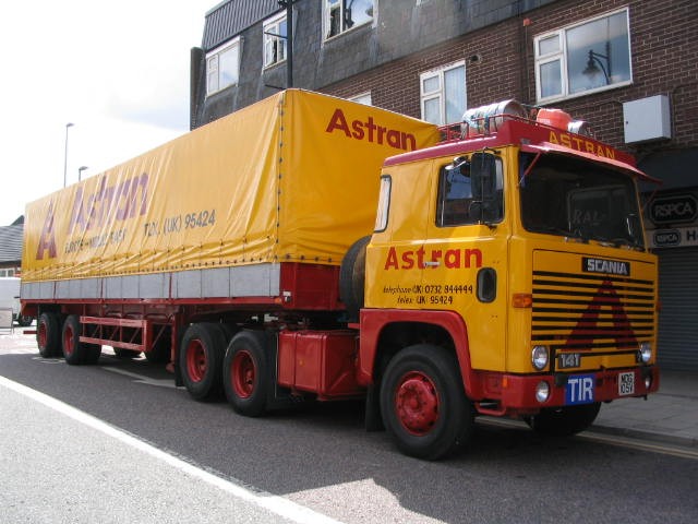 Scania 141 Astrans for wedding transport - Page 1 - Commercial Break - PistonHeads - The image features a large truck parked on the side of a street. The truck is vibrant with a yellow and red striped design, and the word "ASTRAN" is written in red across its side. This cotton candy-colored vehicle appears ready to deliver its goods. The street on which the truck is parked runs along the left side of the image, indicating that the truck is positioned along the curb. In the backdrop, there is a glimpse of a residential area with terraced houses and a brick building, suggesting that the parking spot is near a housing community.